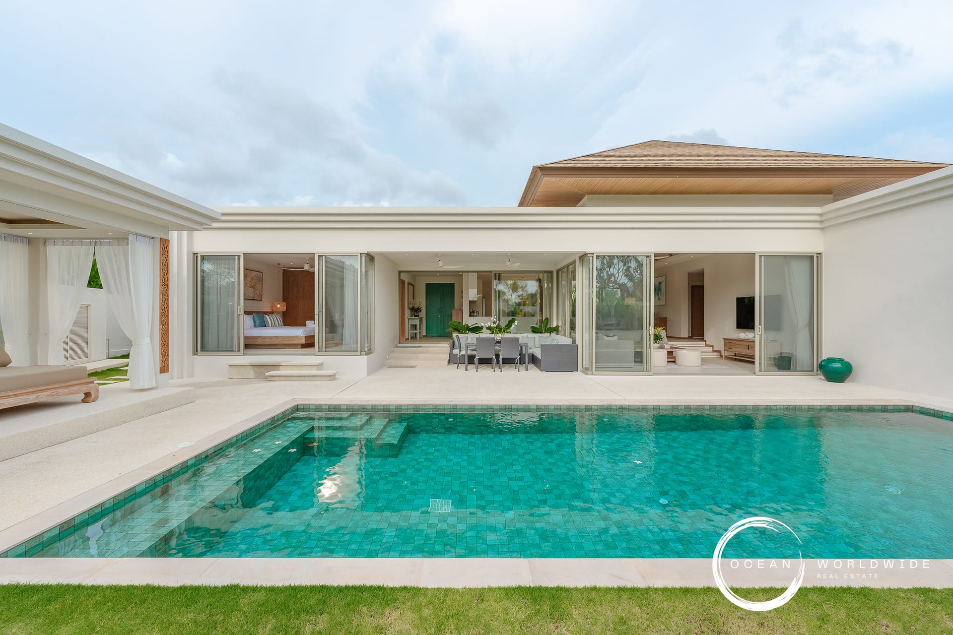 Modern villa with glass sliding doors opening to a courtyard and turquoise pool, outdoor dining area, and green lawn.