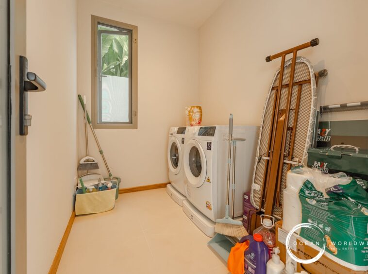 Laundry room with two white front-loading washing machines, ironing board leaned against the wall, and cleaning supplies stacked on the right.