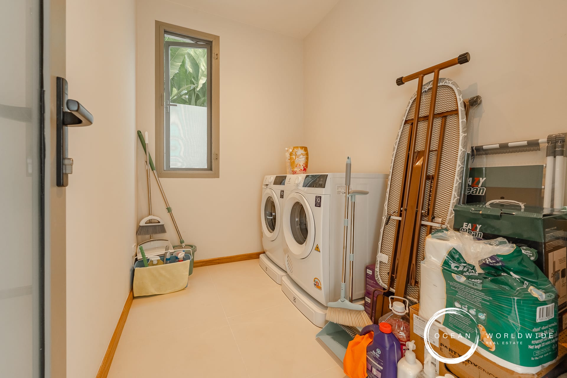 Laundry room with two white front-loading washing machines, ironing board leaned against the wall, and cleaning supplies stacked on the right.